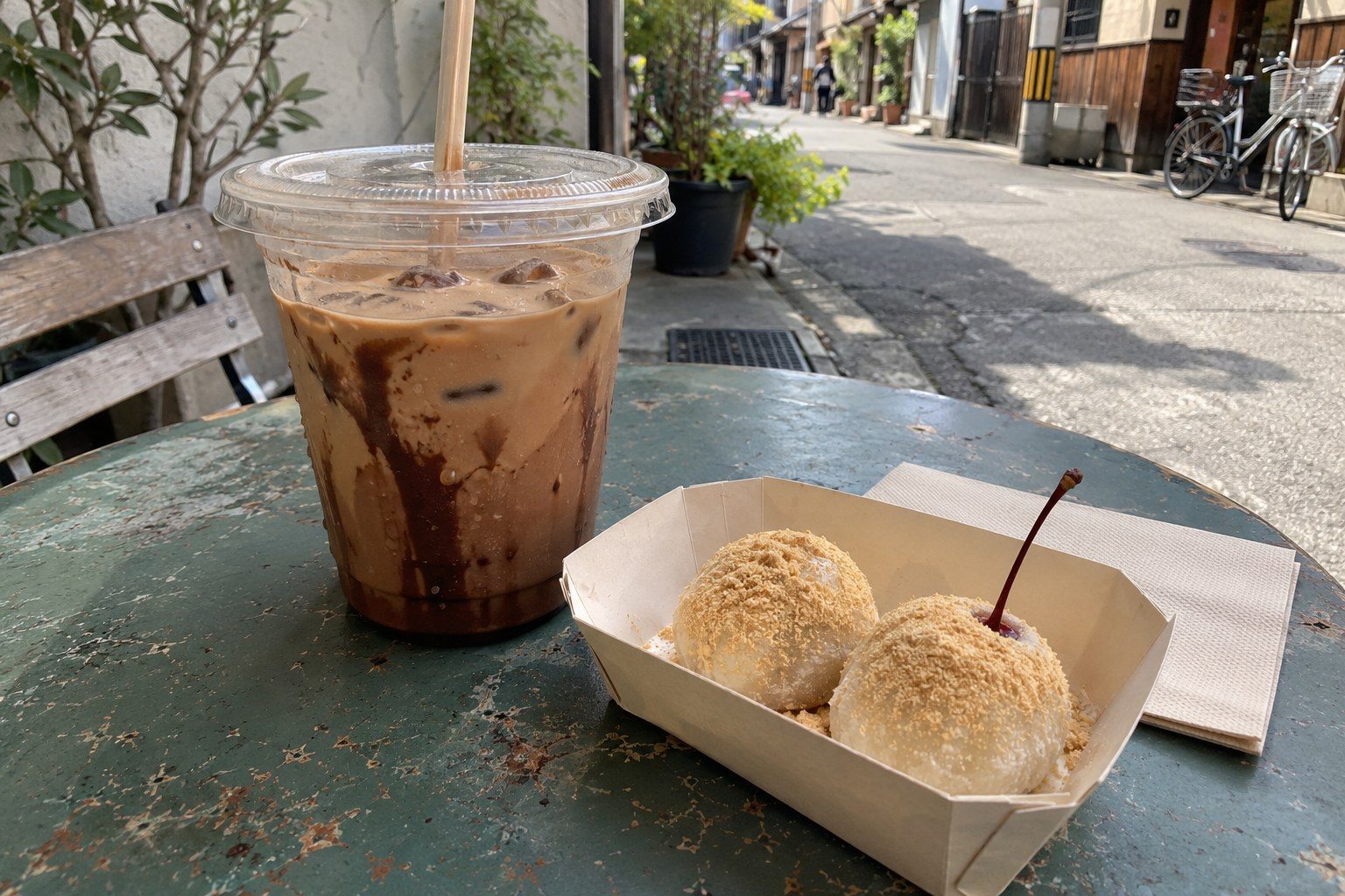 An iced mocha in a plastic cup beside a paper tray of two soybean-dusted mochi, one with a cherry stem, on a café table outside.